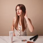 Frau mit glatten, langen Haaren sitzt am Tisch, berührt ihr Haar und hat eine Tube Anti Frizz Creme sowie Stylingwerkzeuge vor sich liegen