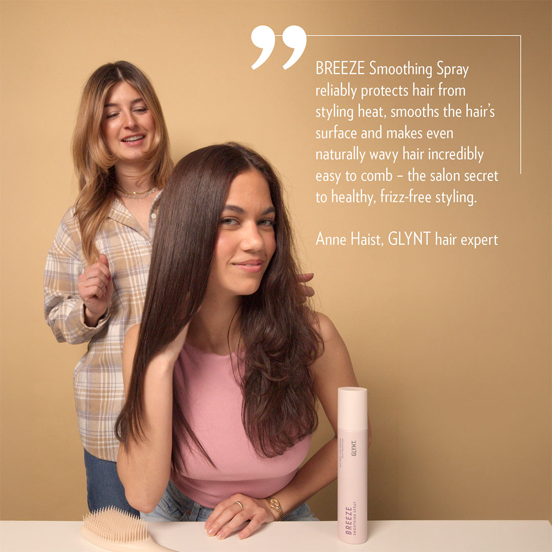 Frau sitzt vor einem Tisch, während eine zweite Frau ihr Haar stylt. Vor ihr steht die Flasche BREEZE Hitzschutz Spray von GLYNT. Das Spray glättet das Haar, schützt vor Styling-Hitze und sorgt für Anti-Frizz-Effekt, sichtbar glattes und glänzendes Haar.