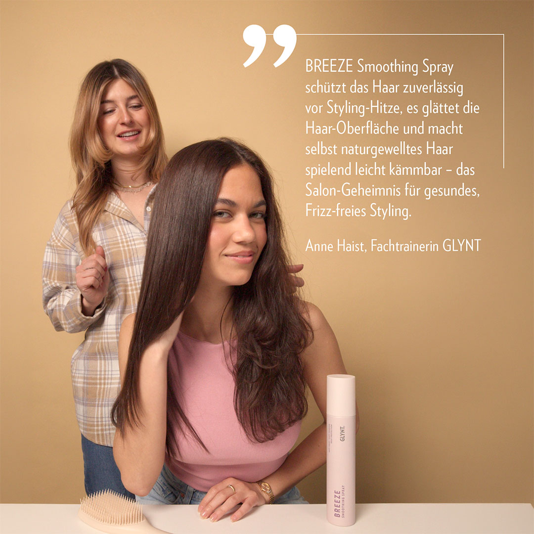 Frau sitzt vor einem Tisch, während eine zweite Frau ihr Haar stylt. Vor ihr steht die Flasche BREEZE Hitzschutz Spray von GLYNT. Das Spray glättet das Haar, schützt vor Styling-Hitze und sorgt für Anti-Frizz-Effekt, sichtbar glattes und glänzendes Haar.