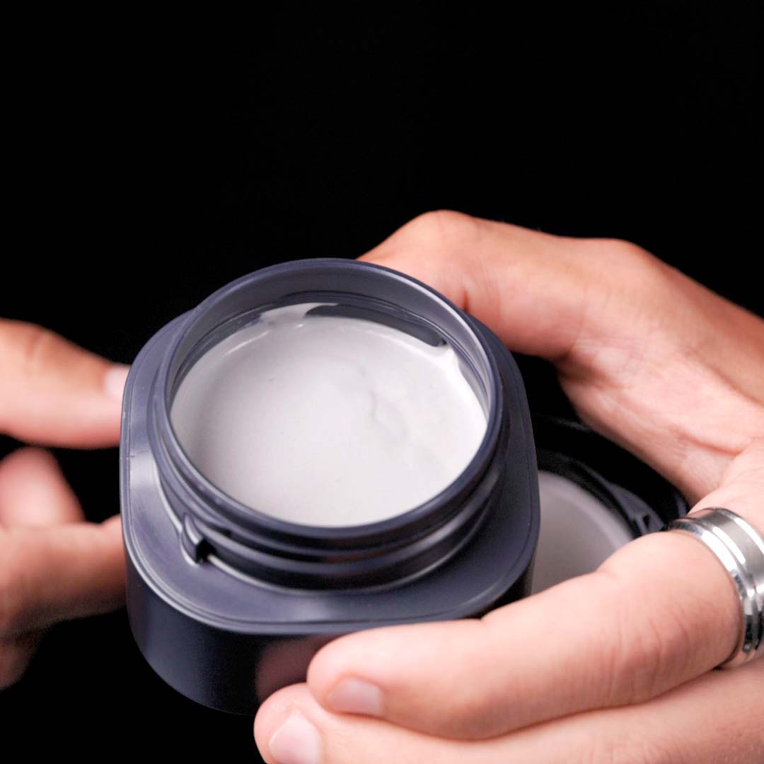 Hochwertige Stylingpaste GRACE Texture Paste von GLYNT in dunkler Dose, geöffnet und in der Hand gehalten. Der Inhalt ist cremig und weiß.