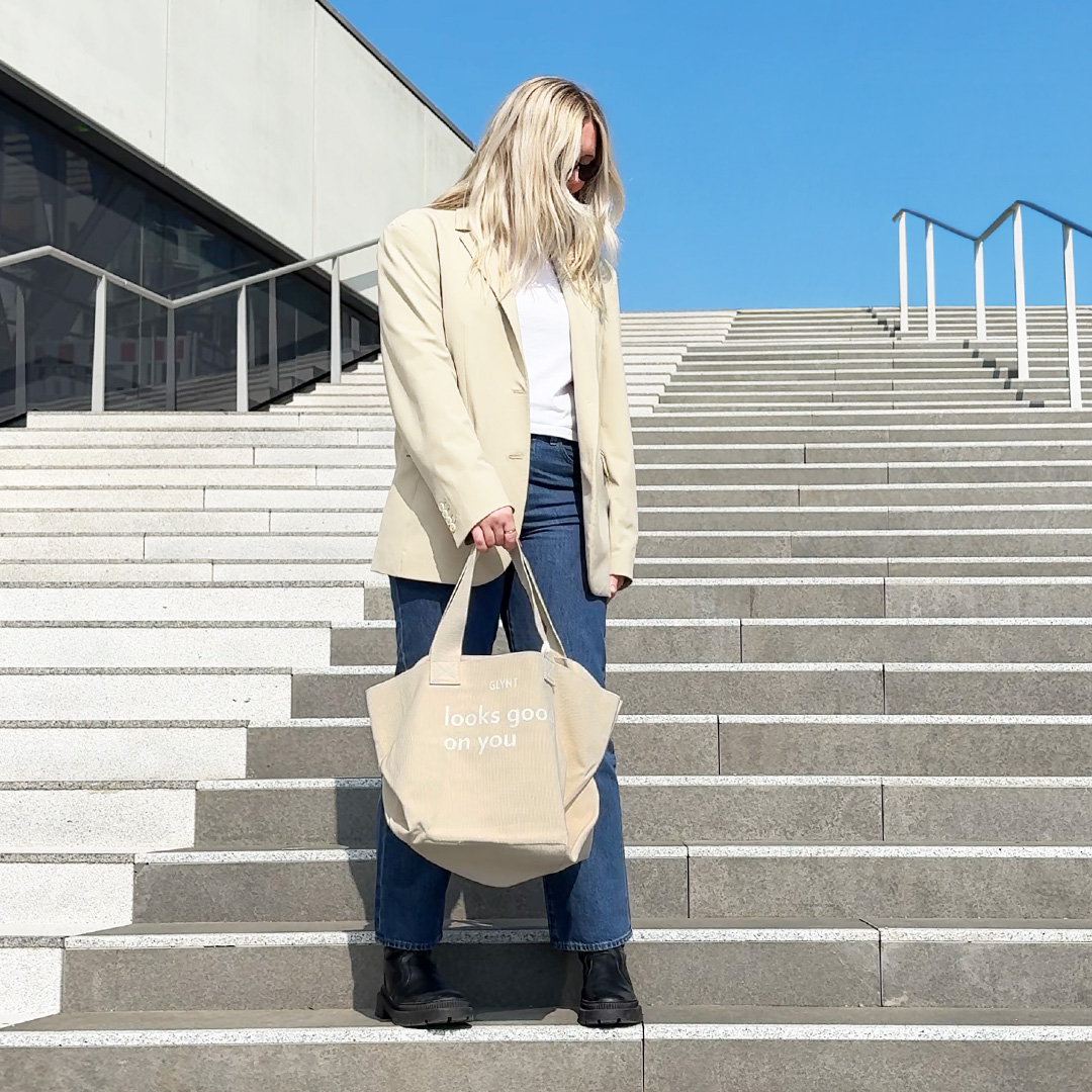 Frau auf Treppe mit beiger GLYNT Stofftasche „looks good on you“ in urbaner Umgebung.